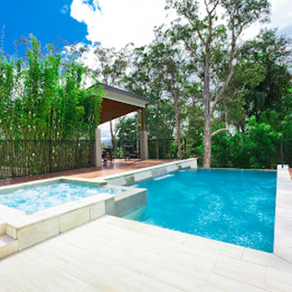 Piscina e spa integrado cercados por deck de pedra clara e área de estar coberta