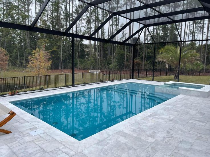 Piscina con deck de travertino blanco y enclosure negro enmarcando los árboles